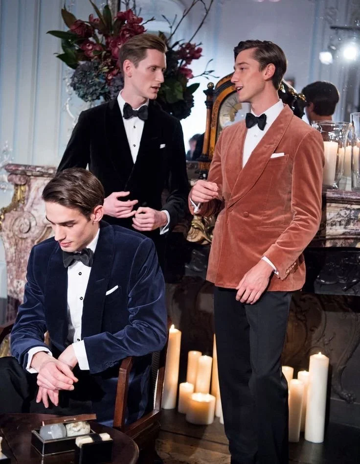Three men in formal tuxedos and suits, engaging in conversation in an elegant room with candles, flowers, and a vintage clock in the background.