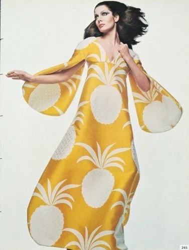 A woman wearing a long yellow dress with white pineapple and leaf patterns, posing with her arms outstretched.