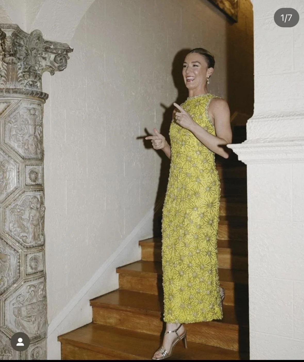 A woman in a yellow, beaded, sleeveless dress standing on a wooden staircase, smiling and pointing with both hands.