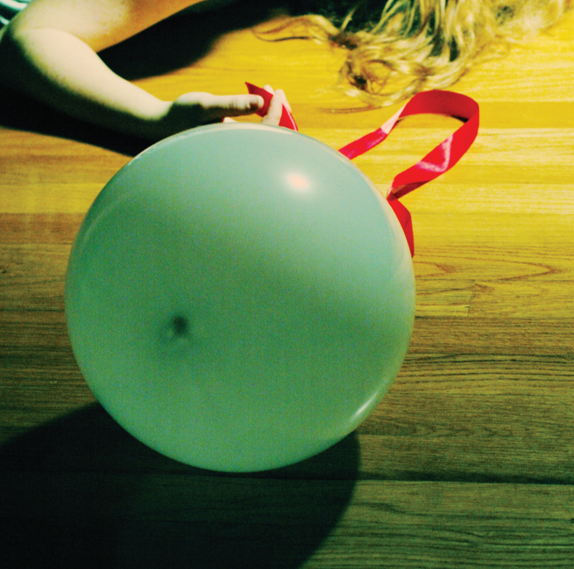 A person with long hair lying on a wooden floor holding a green balloon with a red ribbon tied to it.