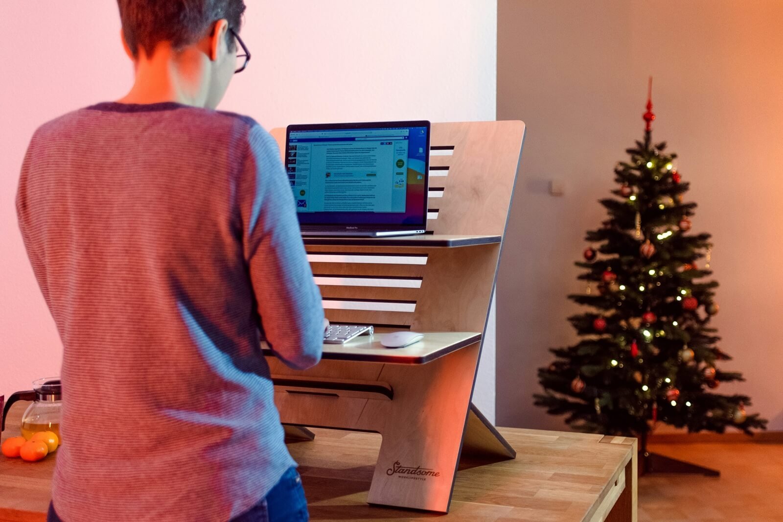 A person with glasses wearing a red and gray sweater is standing in front of a computer standing desk with a black MacBook laptop open. A small Christmas tree decorated with ornaments and lights is in the background.