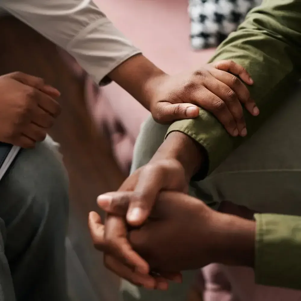 Two people holding hands in a supportive manner, one resting their hand on the other's arm in a comforting gesture.