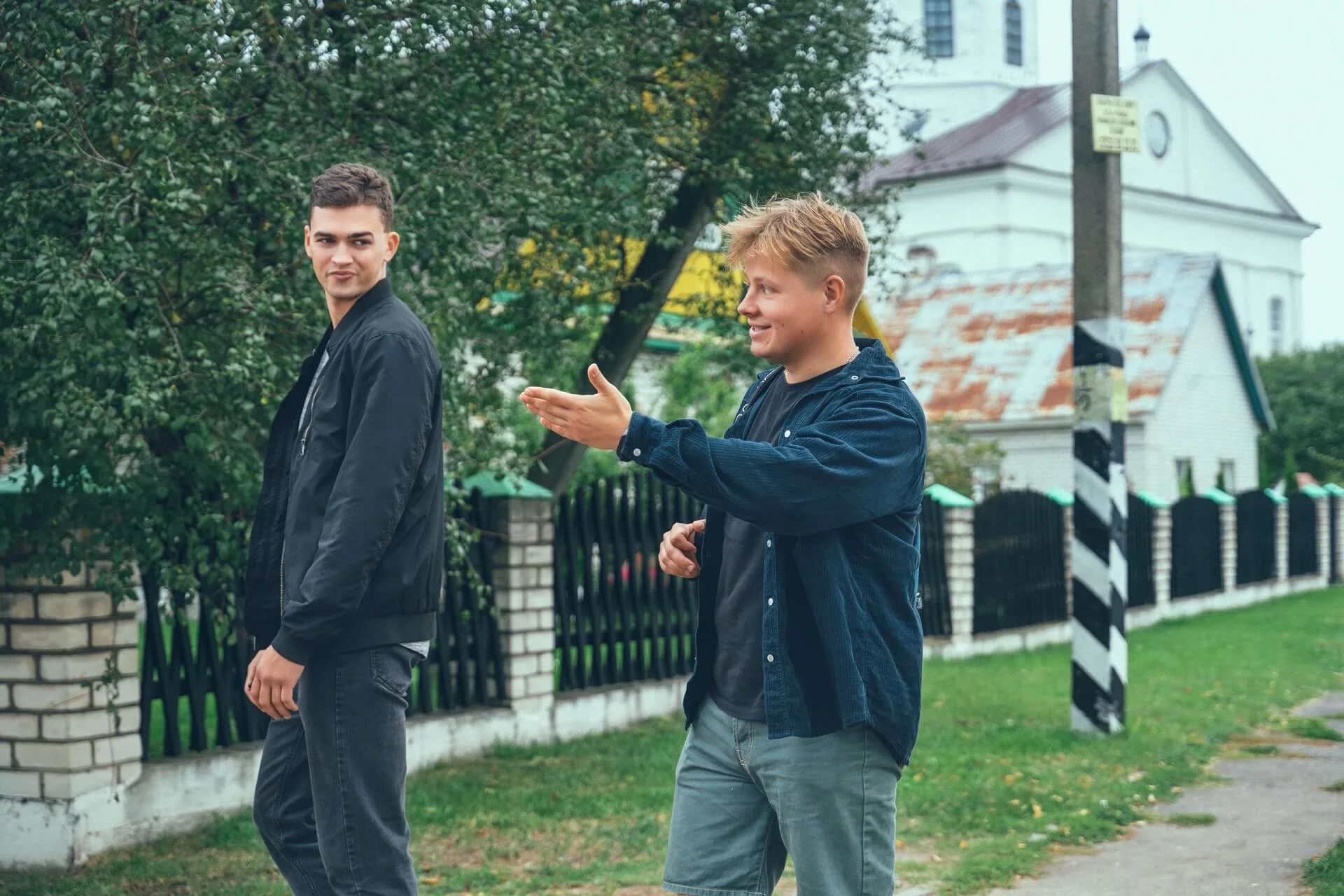Two young men, one with dark hair and wearing a black jacket, and the other with light hair and wearing a dark shirt, engaging in a conversation outdoors near a painted crosswalk barrier, with trees and a white building in the background.