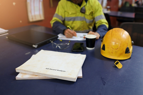 Construction worker wearing safety gear writing at a desk with plans, a laptop, a cup, and a yellow helmet.