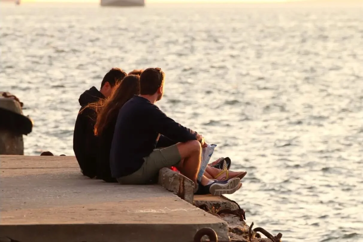 Four young people sitting on a concrete dock by the water at sunset, facing away from the camera and looking at the sea.