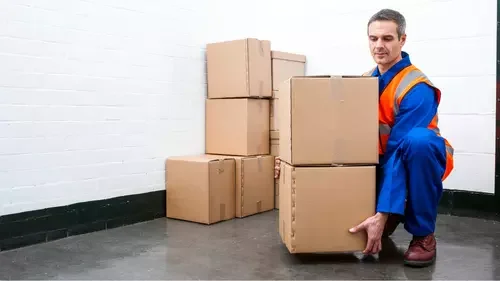 A worker in a safety vest and blue uniform is handling a cardboard box in a storage room with white walls and black floor baseboard.