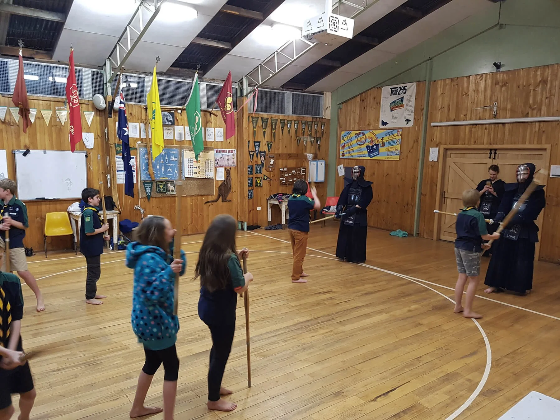 A group of children participating in a martial arts class inside a gymnasium with wooden floors and walls. They are practicing sword techniques under the supervision of two individuals dressed in traditional kendo armor. The gym has various flags, posters, and decorations on the walls.
