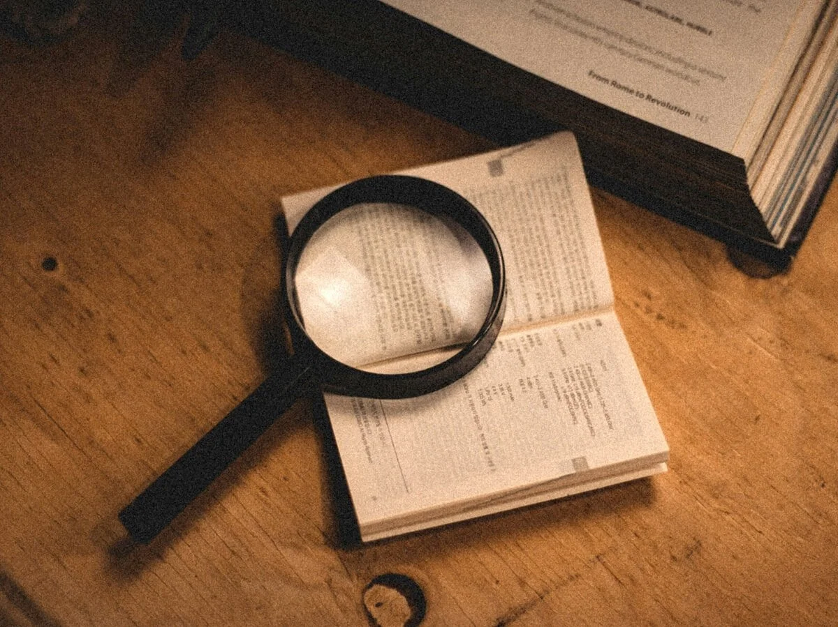 An open book with a magnifying glass on top, on a wooden surface, with a closed book nearby.