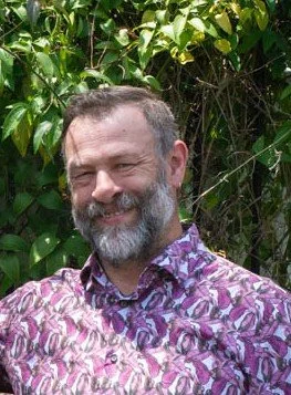 A man with a beard and short hair smiling outdoors in front of green foliage, wearing a purple patterned shirt.