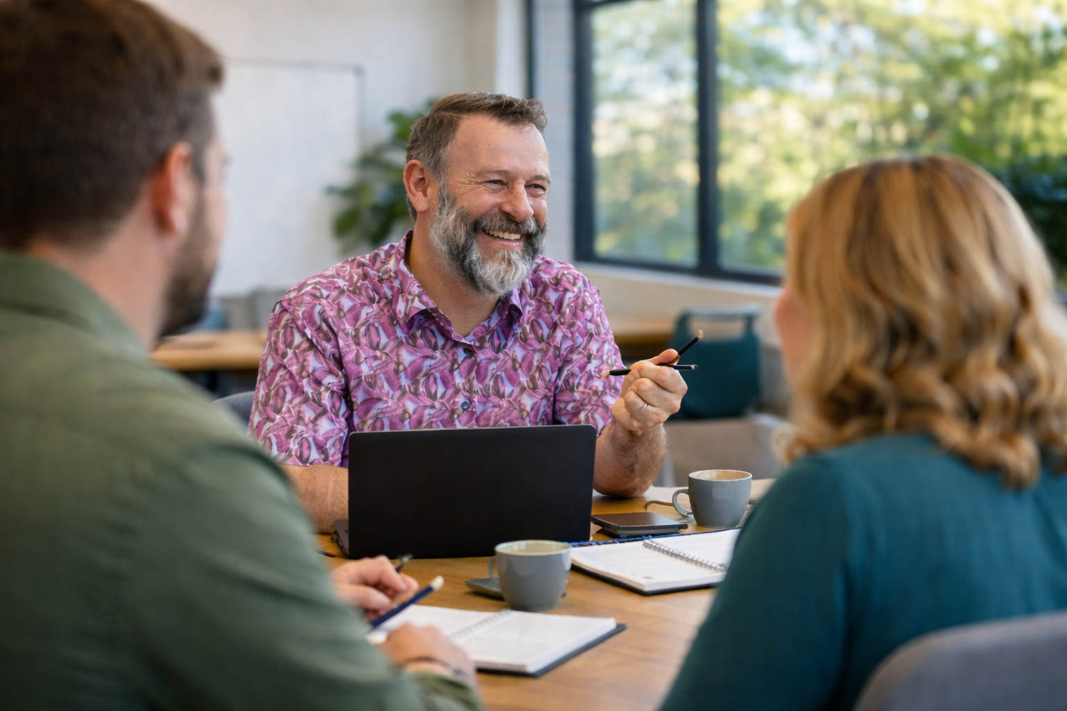 Three people in a meeting room having a discussion, with notebooks, a tablet, and coffee cups on the table, a man with a beard and purple patterned shirt smiling.