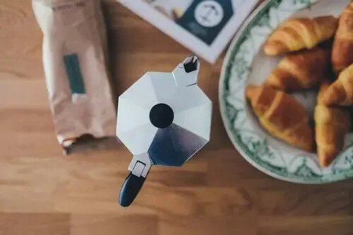 A top view of a coffee maker, a plate of croissants, and a paper bag on a wooden table.