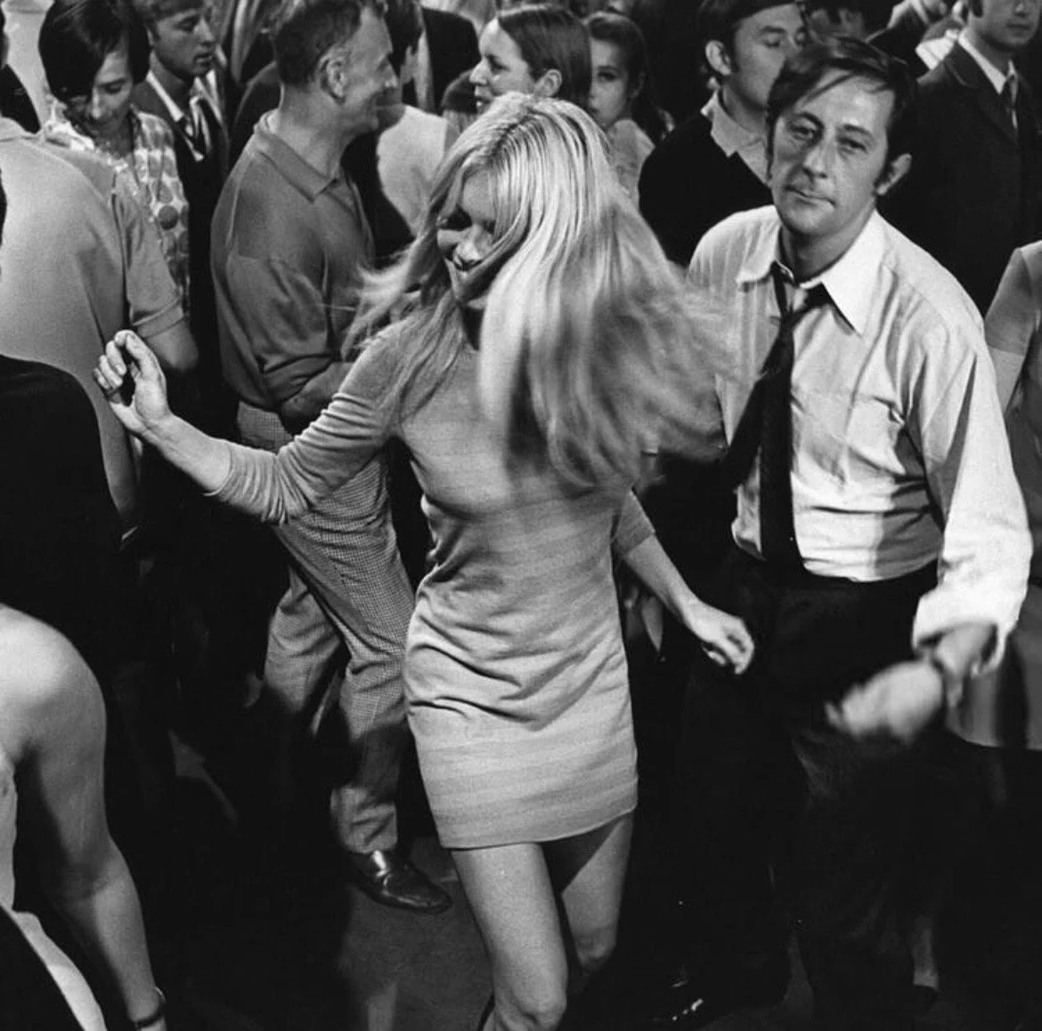 A black-and-white photo of a woman with long blonde hair dancing with a man at a crowded party or dance event. The woman is wearing a form-fitting dress, and the man has a white shirt with a loosened tie. They are holding hands, surrounded by other people who are also dancing or socializing.