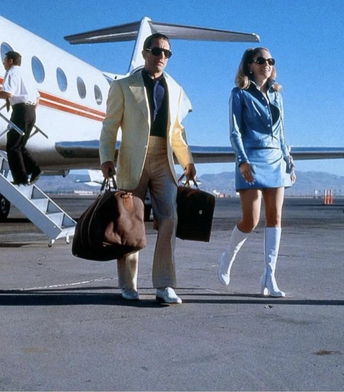 A man and woman walking on an airport tarmac away from a private jet, carrying luggage and dressed in stylish, retro clothing.
