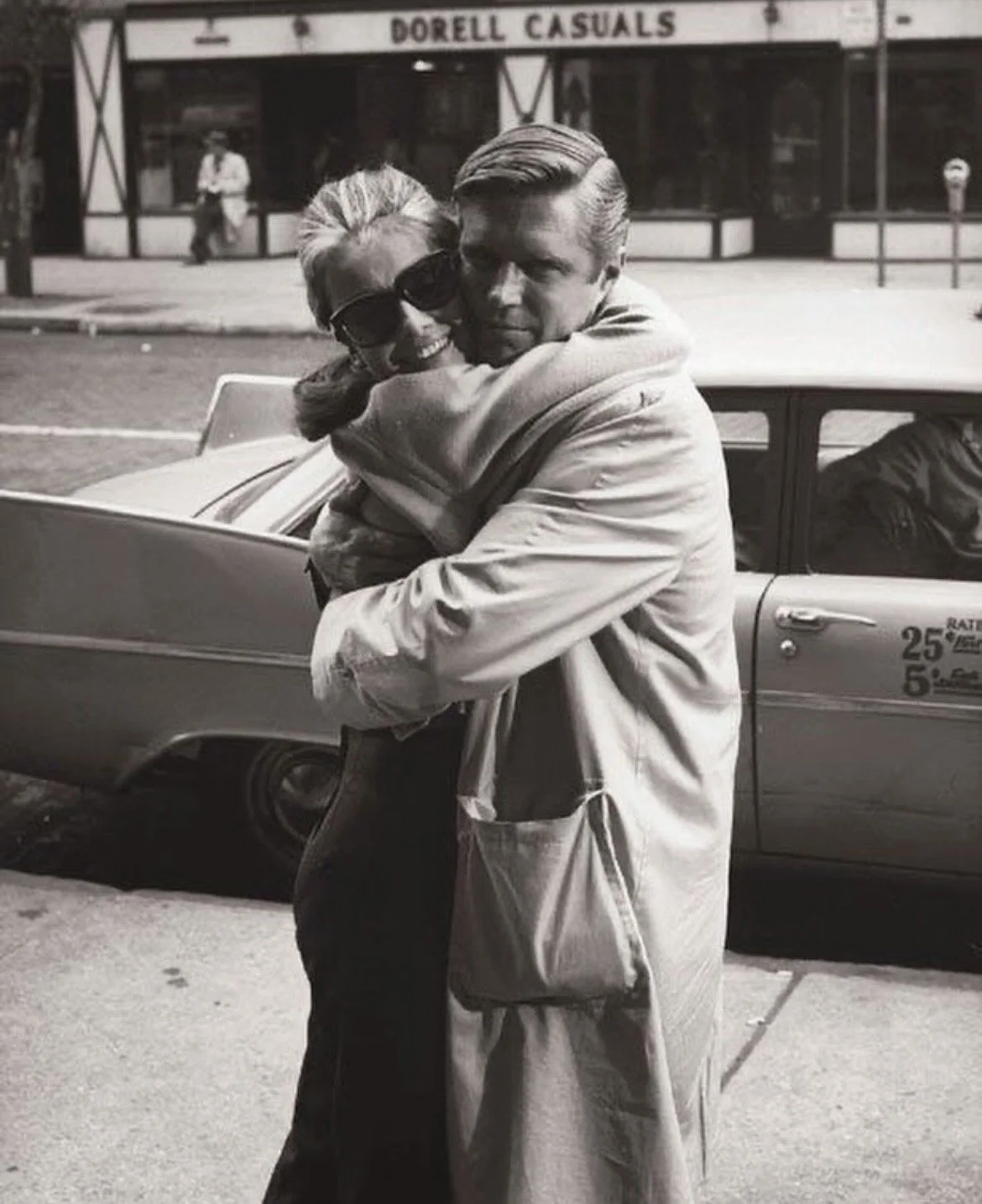 A black-and-white photo of a man and woman hugging on a city sidewalk. The woman is wearing sunglasses and smiling, while the man is looking directly at the camera with a serious expression. A vintage car is parked behind them, and a storefront with the sign "Dorell Casuals" is visible in the background.