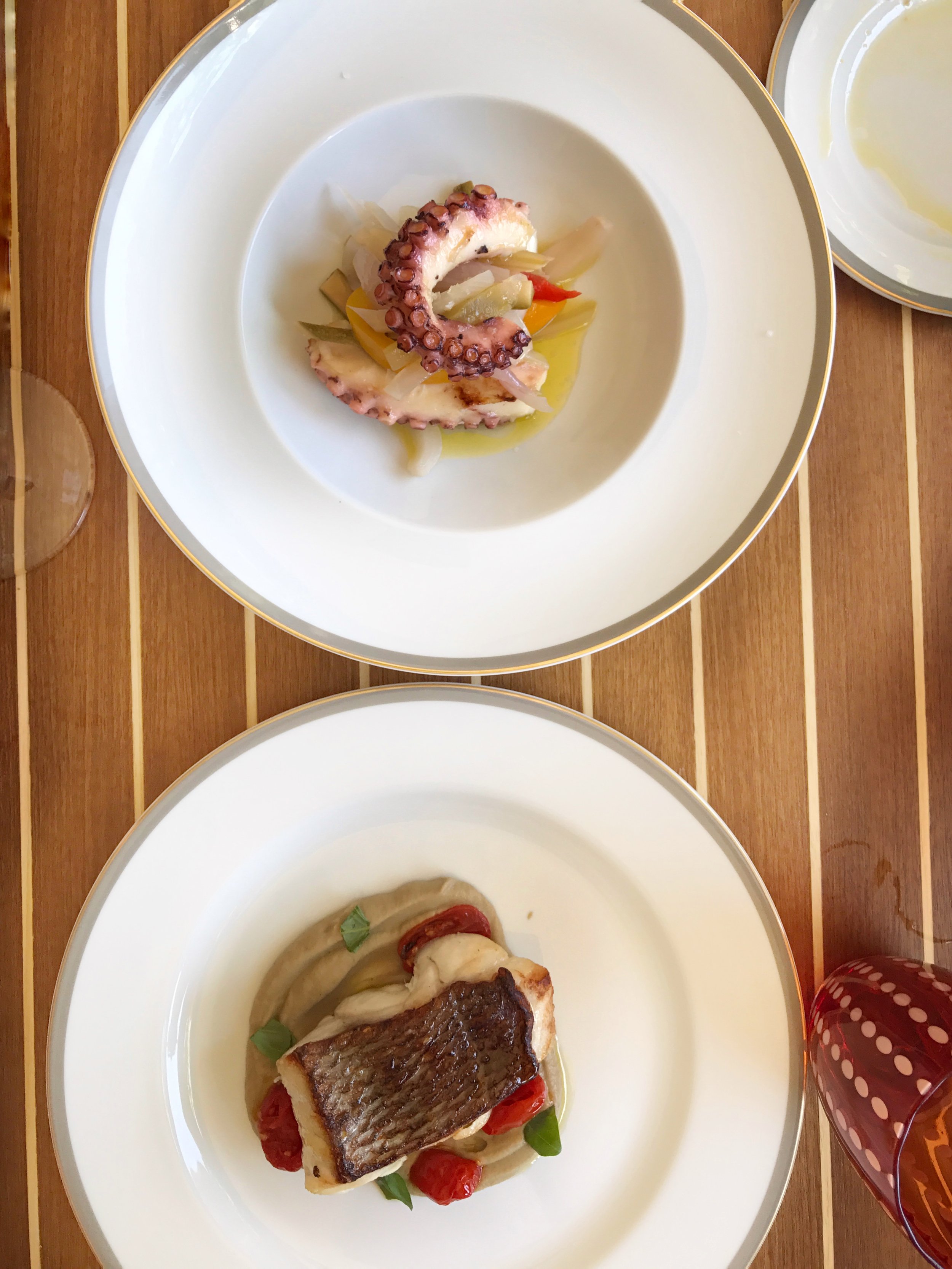Two plated gourmet dishes, one featuring octopus tentacle with vegetables, the other a piece of grilled fish with cherry tomatoes and greens, on a wooden table.