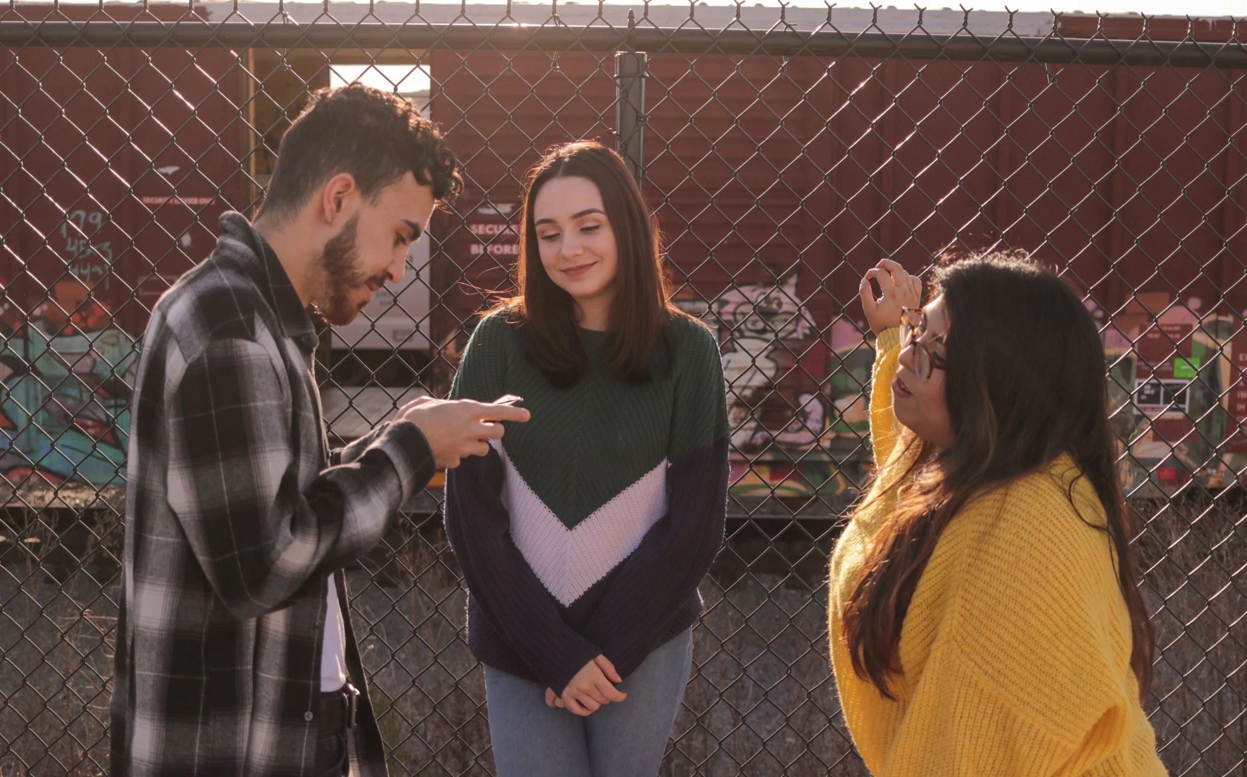 Three young women standing behind a chain-link fence, one is smiling while looking at her phone, another is leaning against the fence with her hand, and the third is looking at her phone with focused attention. They are outdoors with sunlight and a red structure with signs in the background.