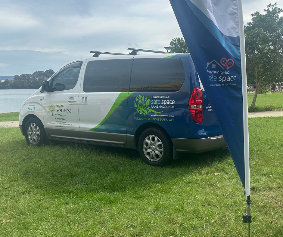 A white and blue van parked on grass near a lake with trees in the background, featuring logos and text promoting community-led safe space and suicide prevention initiatives.