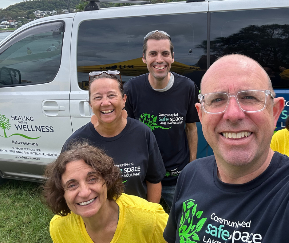 Four smiling people posing outdoors next to a vehicle with wellness and mental health support branding.