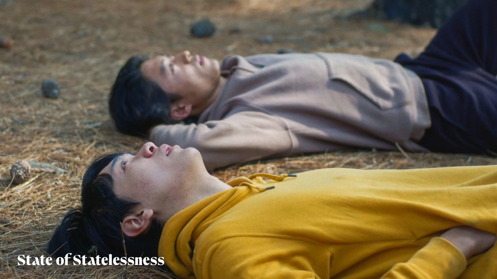Two young men laying on the ground outdoors, looking up into the sky. One wears a yellow hoodie and the other a beige hoodie. The ground is covered with dry pine needles and small rocks.