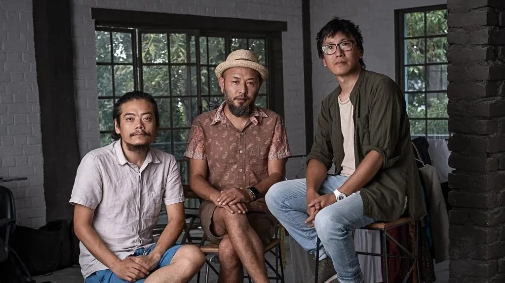 Three men sitting indoors in front of large windows with greenery outside, casual clothing, relaxed pose.