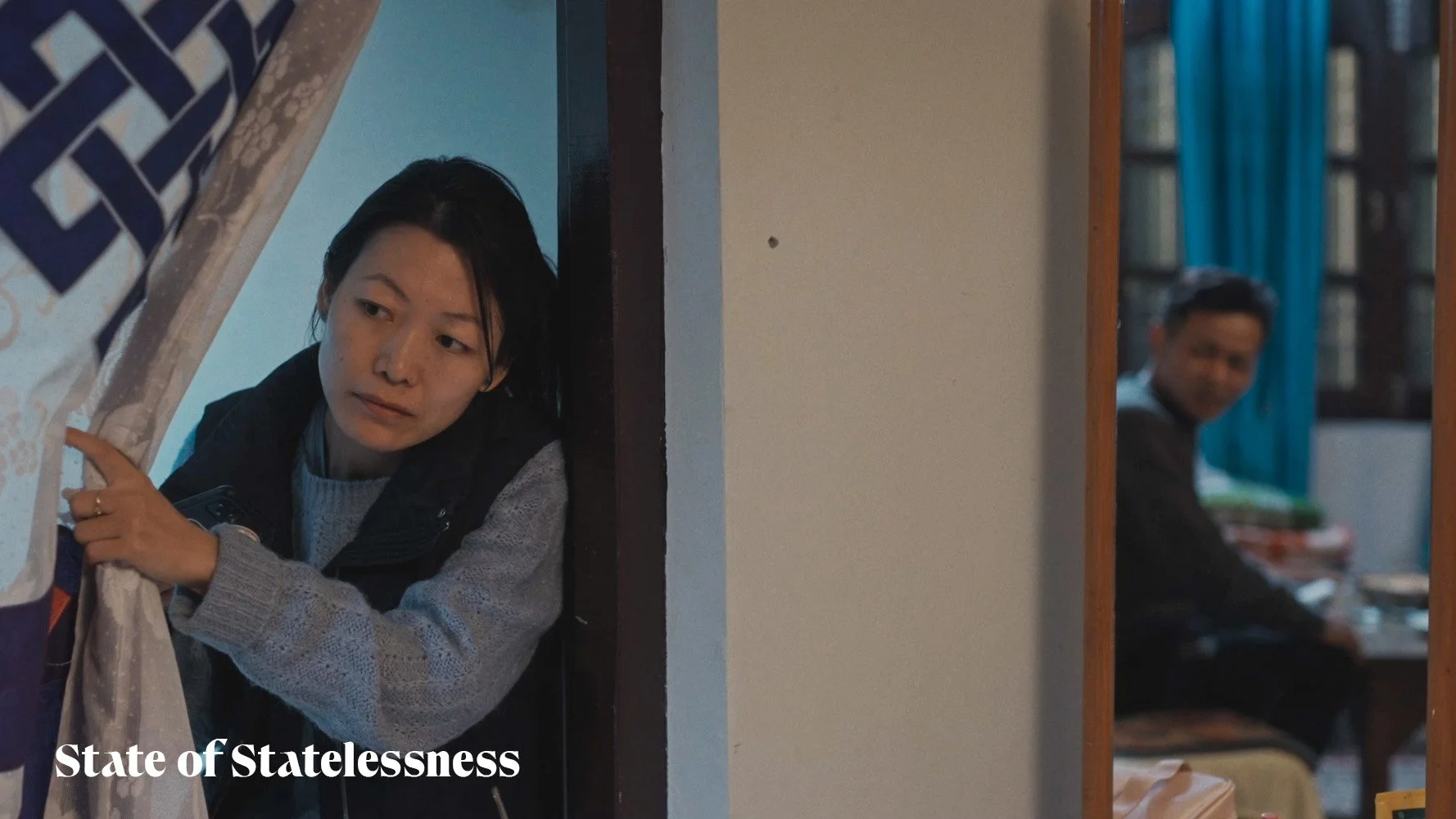 A woman peering around a partially open curtain, with a man sitting at a desk in the background, in a dimly lit room.
