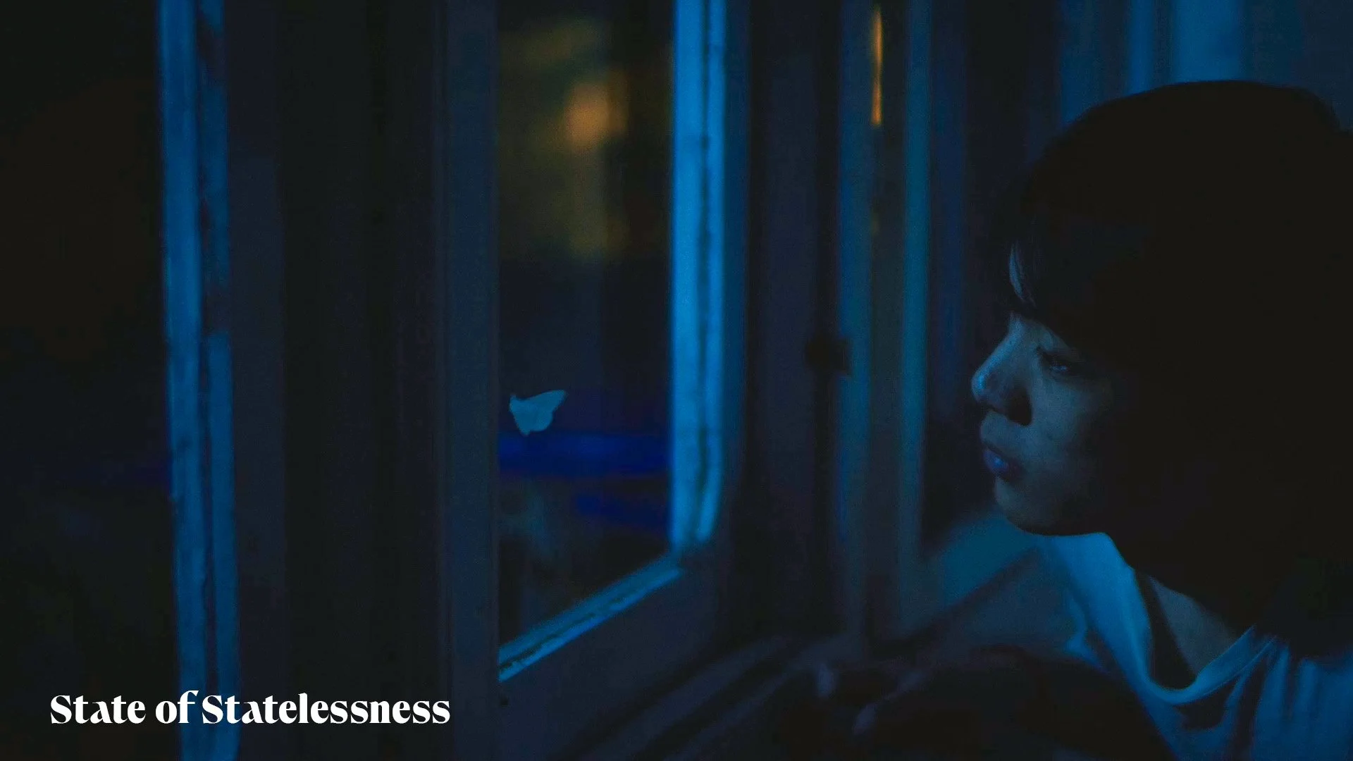 Side profile of a young person looking out of a window at night, with a dark background and blue lighting.