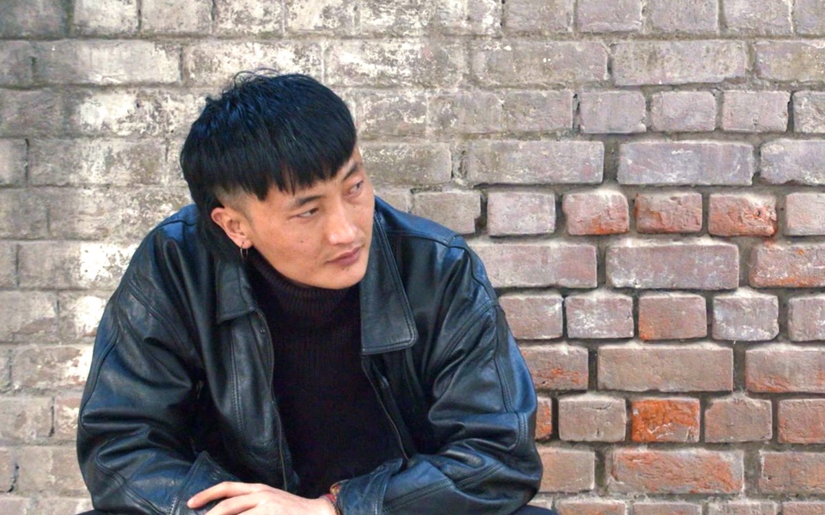 A young man with black hair, wearing a black leather jacket and black turtleneck, sitting in front of a brick wall, looking to the side.