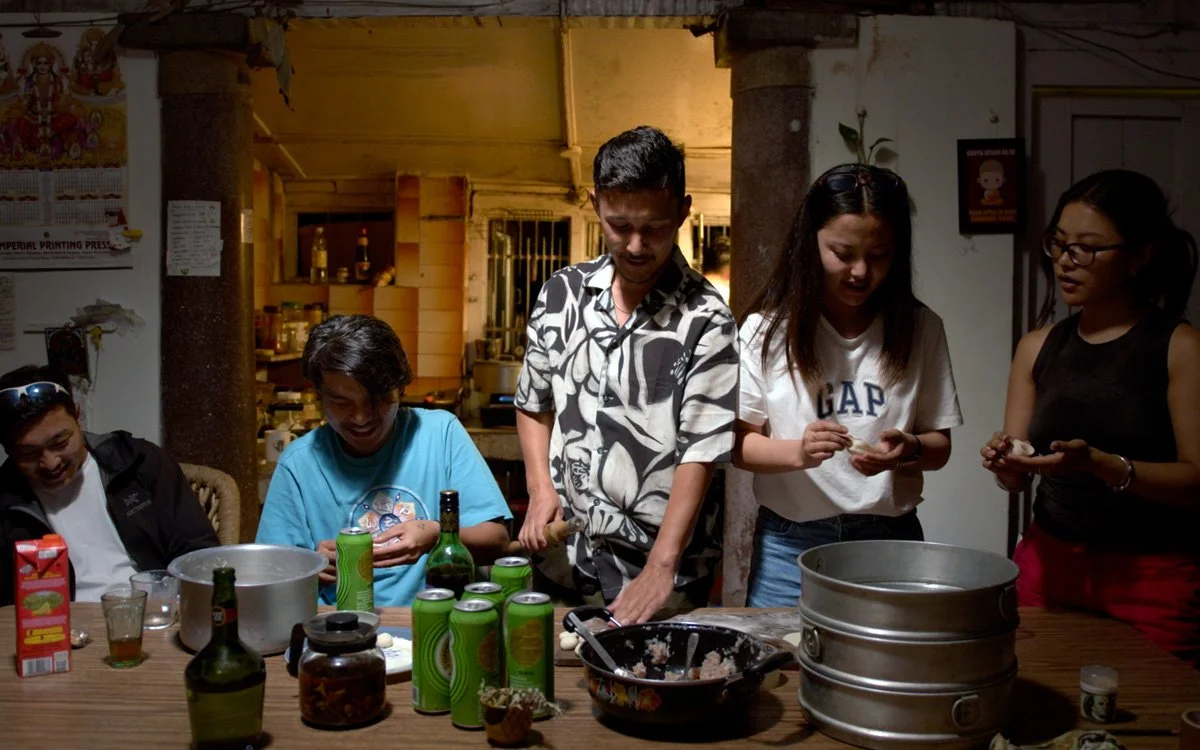 Five people gathered around a table in a cozy indoor setting, some preparing food while others are laughing and chatting. The table features bottles, cans, and cooking utensils.