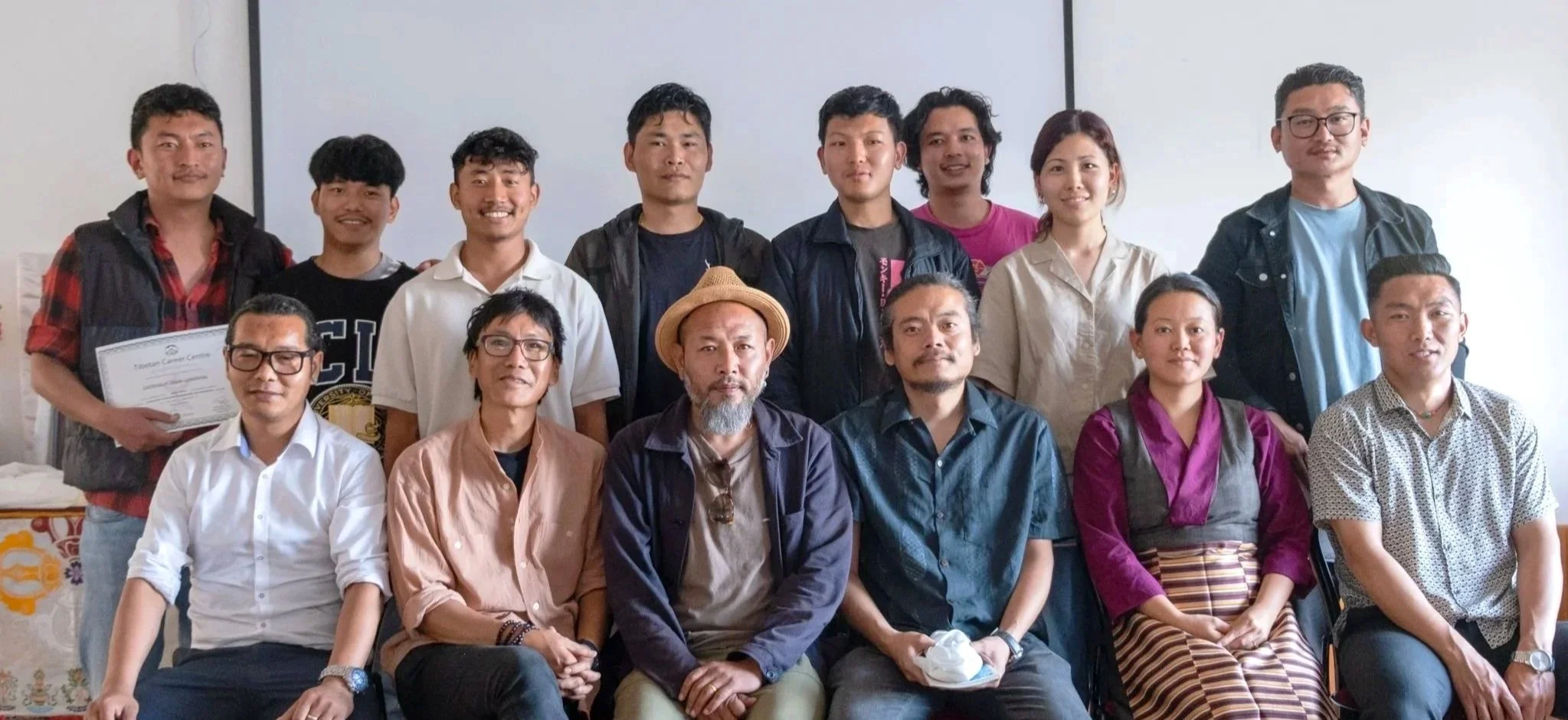 Group photo of 15 diverse people, some seated and some standing, indoors in front of a plain white wall. Some are smiling, wearing casual and business casual attire, and one person holds a certificate.