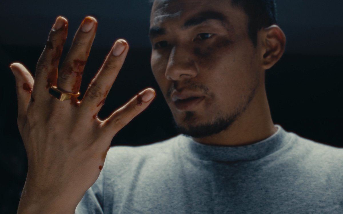 A young man with blood on his fingers and face, looking at his hand with a serious expression, wearing a gray shirt.