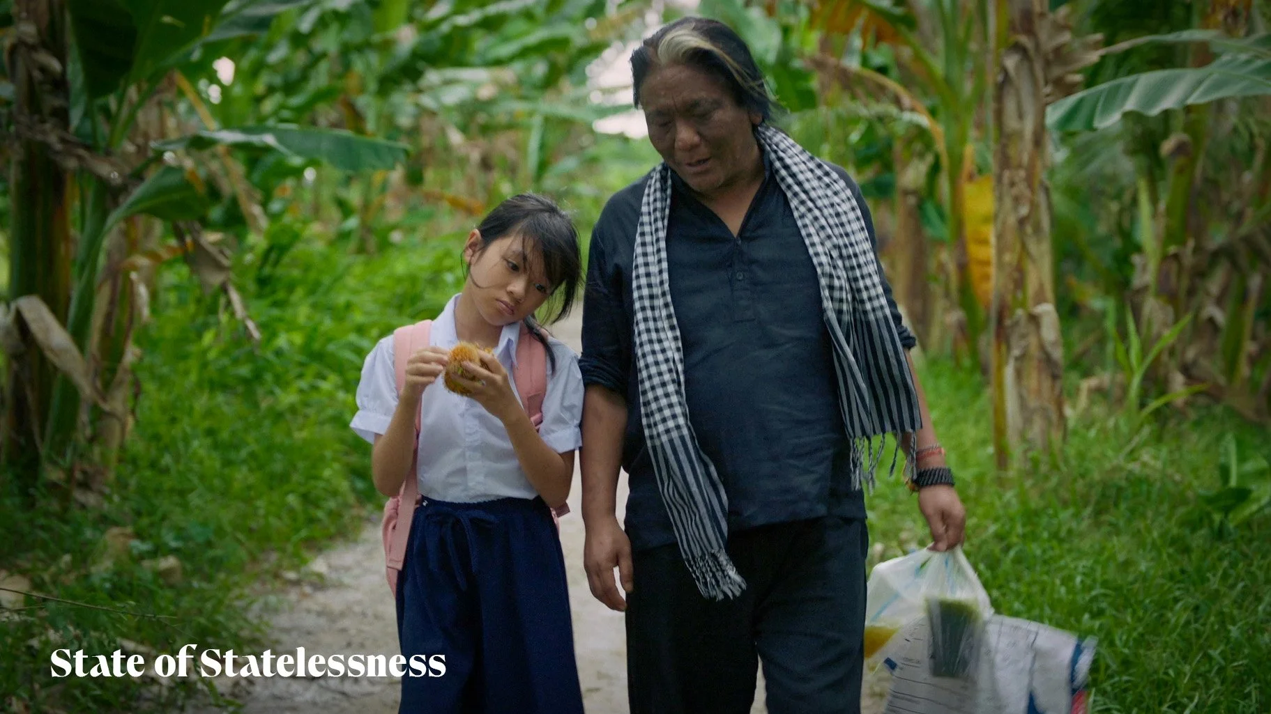 A young girl in a school uniform walking with an older man through a lush, green cornfield. The girl is holding a piece of food, and the man carries a plastic bag. They appear to be having a conversation.