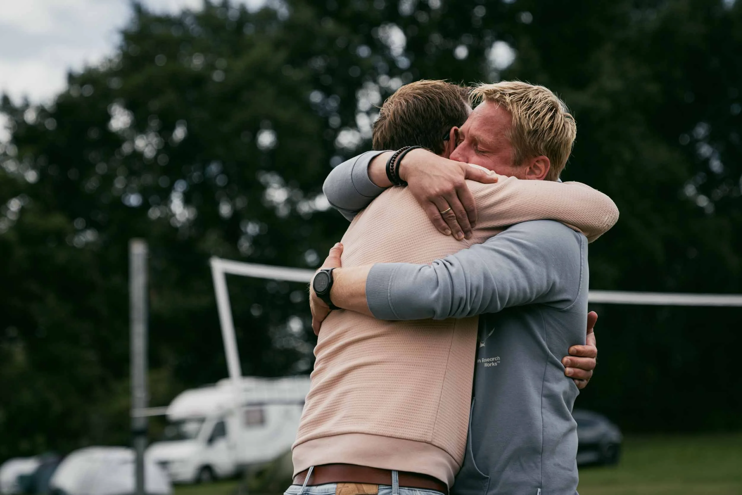 Two men hugging outdoors with cars and trees in the background.