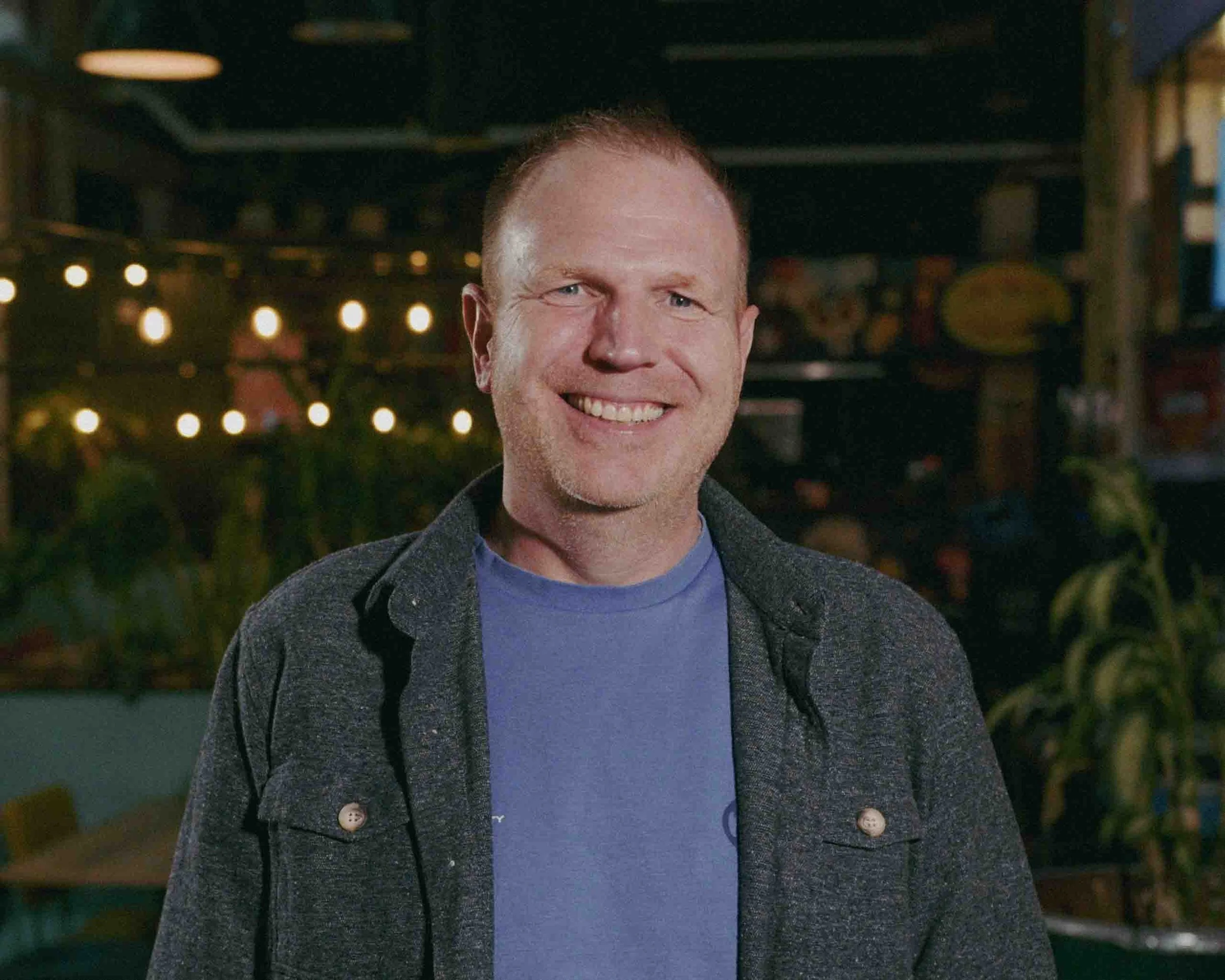 Two men sitting and talking in an indoor setting, one man with a colorful hoodie and the other with a dark t-shirt. The man in the hoodie has a beard and is gesturing with his hand on his chest, while the other man is smiling and sitting crossed-legged.