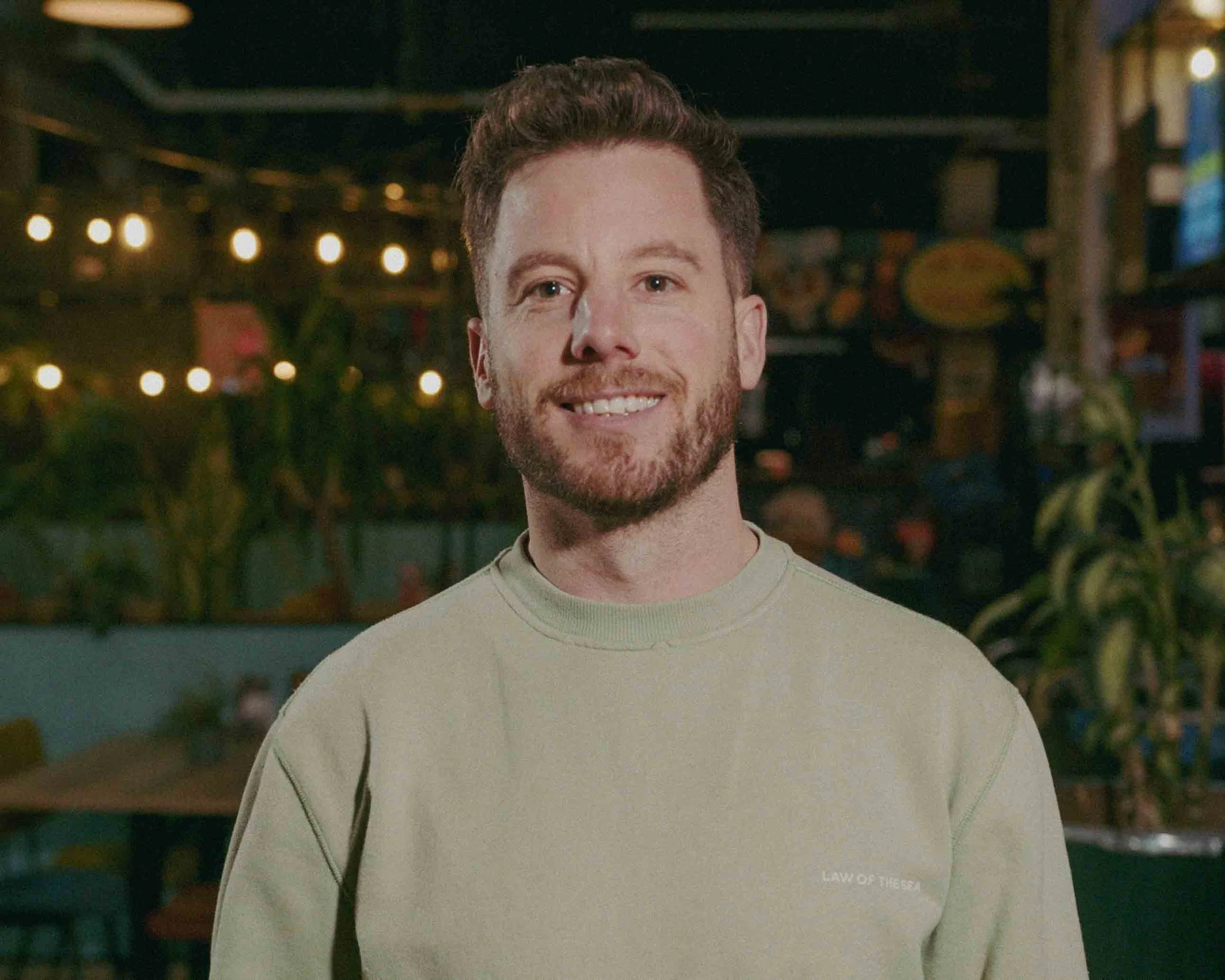 A man relaxing in a hot tub outdoors with five people standing around him, some holding drinks, on a cloudy day.