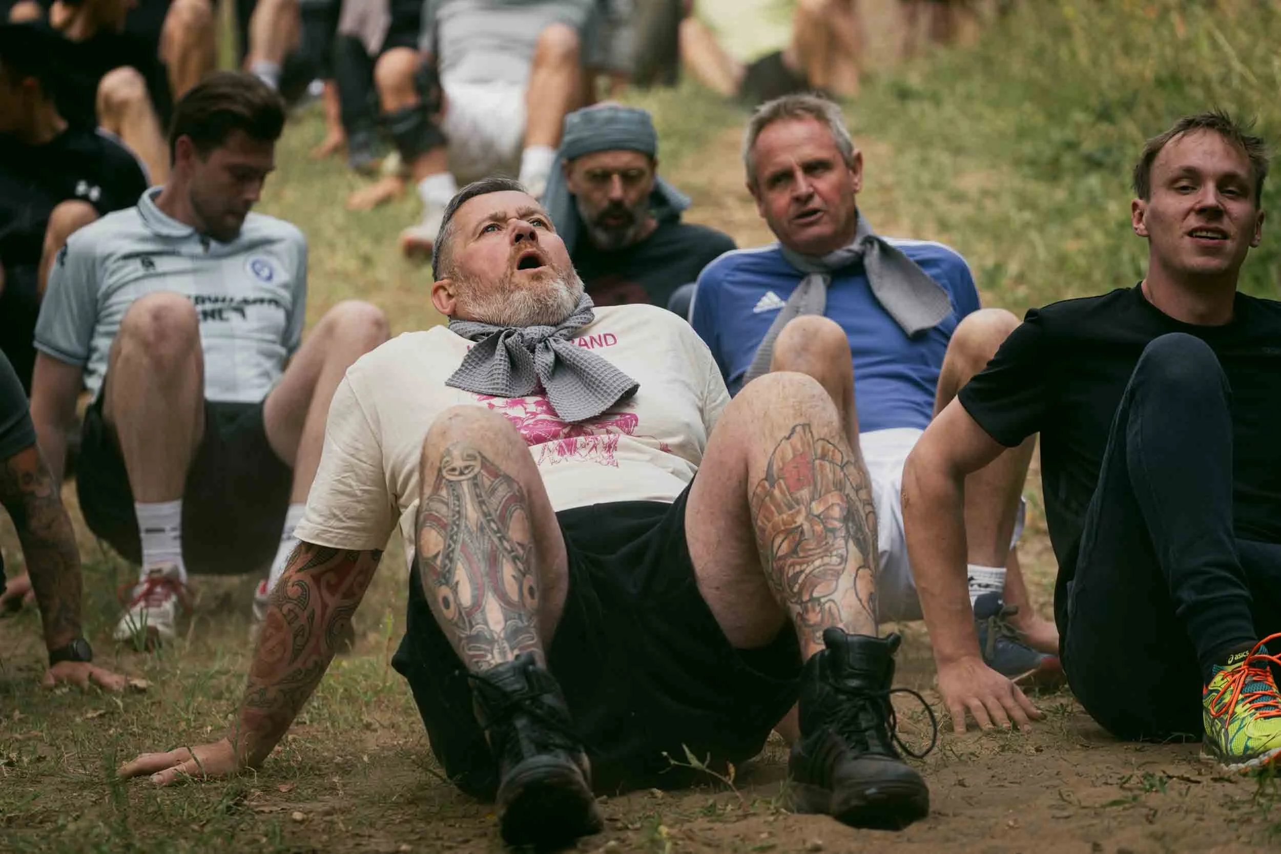 A group of men participating in a challenging outdoor activity, with some sitting on the ground and one man with tattoos on his legs and arms in the foreground, wearing a beige t-shirt and black shorts, looking up with an intense expression.