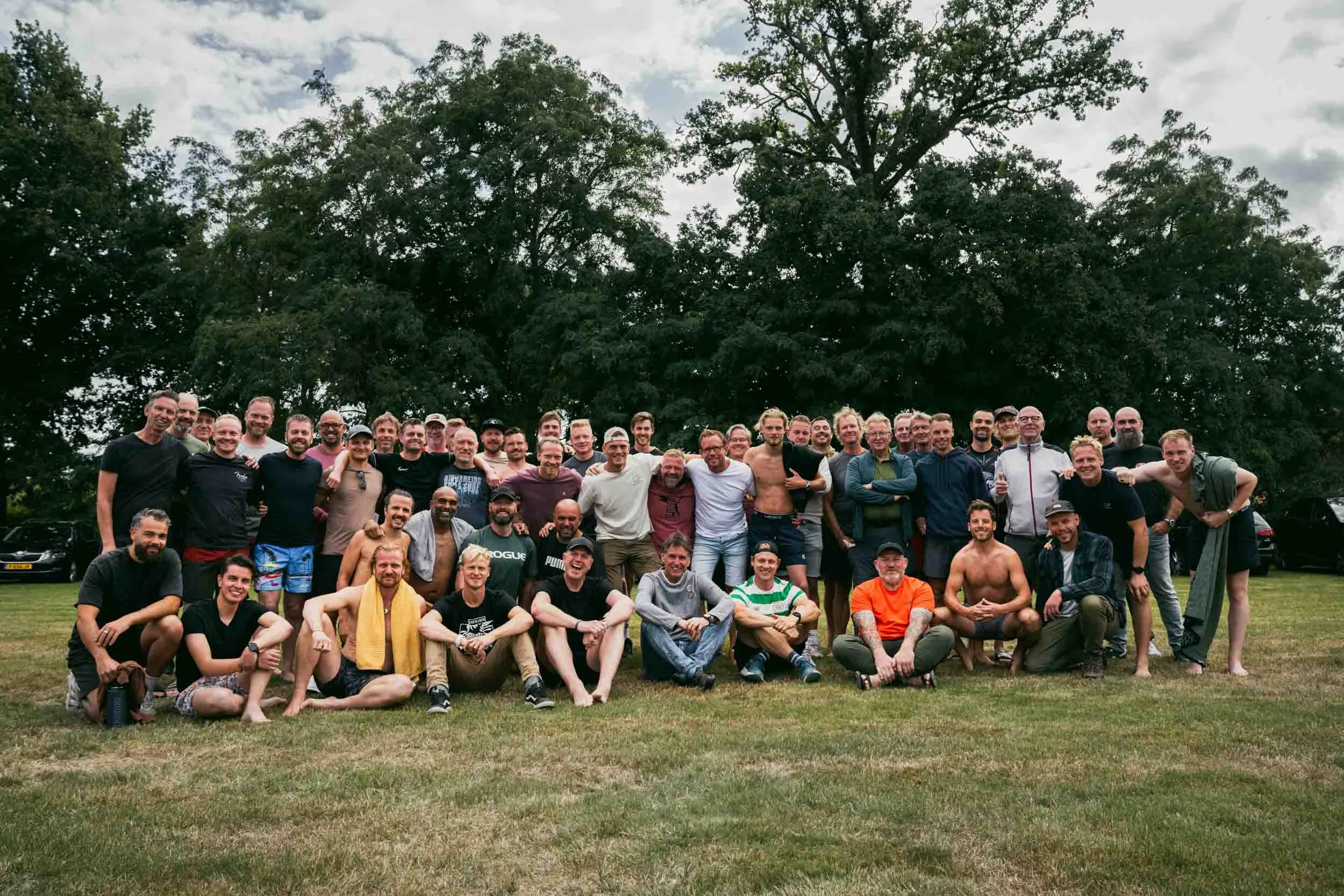 A large group of men gathered outdoors on a grassy field in front of trees, posing for a group photo, some standing and some sitting on the ground, under a cloudy sky.
