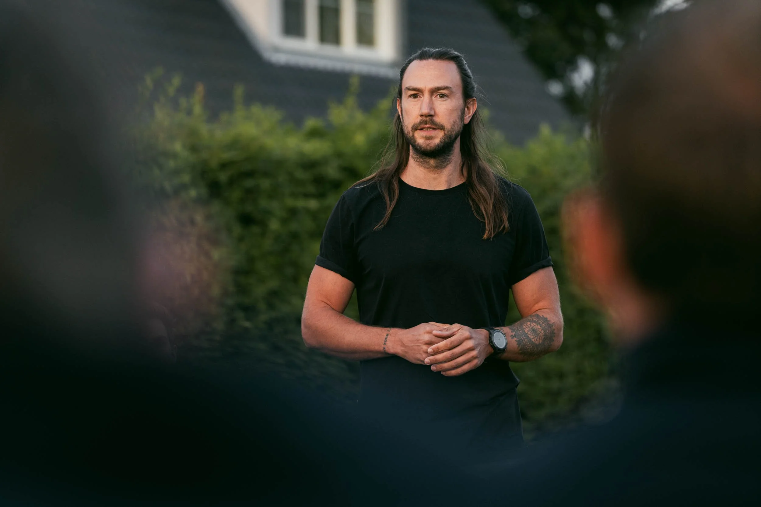 Man with long dark hair, beard, wearing a black t-shirt and smartwatch, standing outdoors with greenery in the background.