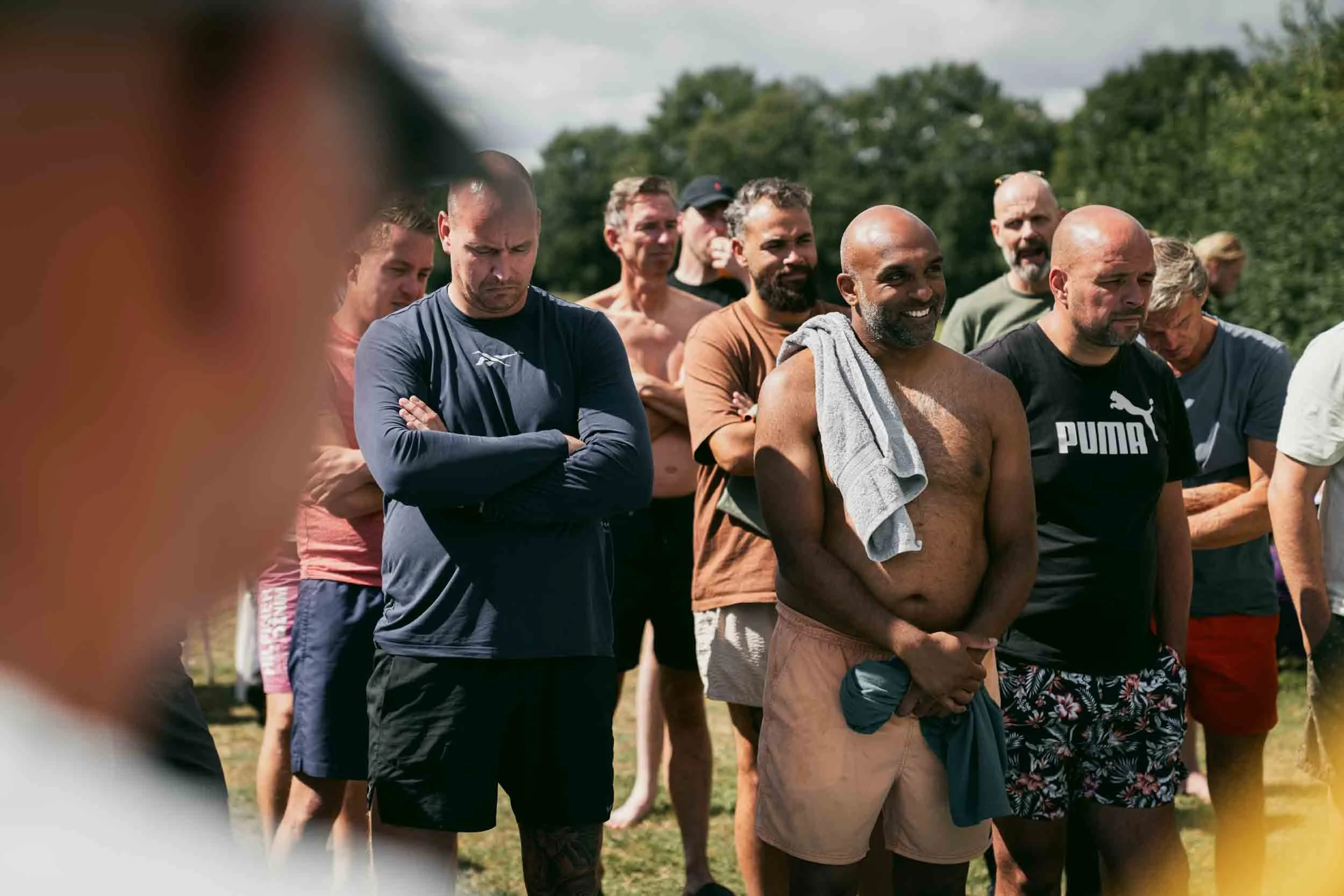 Group of men, some shirtless, gathered outdoors on a grassy field, listening attentively.
