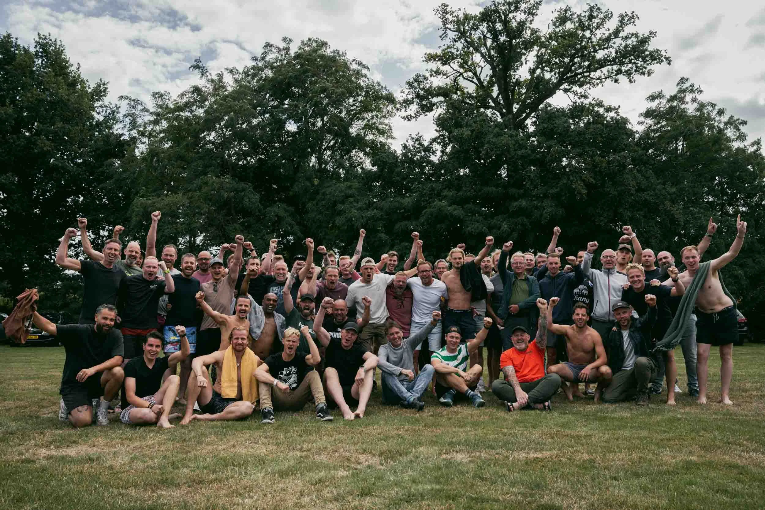 Large group of people outdoors celebrating, some with shirtless and others with casual clothing, raising their fists, standing and sitting on grass, with trees and cloudy sky in the background.