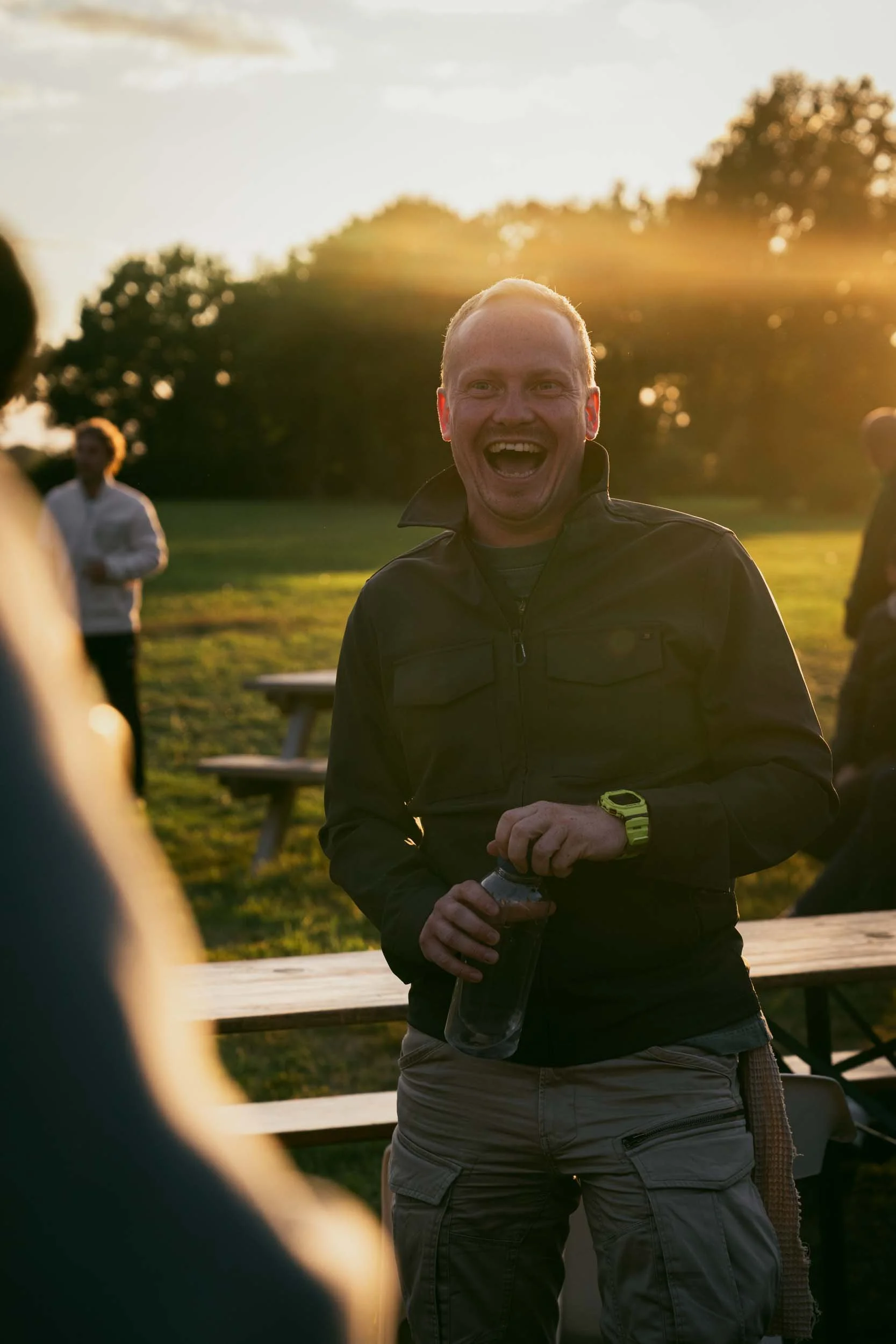 A man smiling and holding a water bottle outdoors during sunset, with other people and picnic tables in the background.