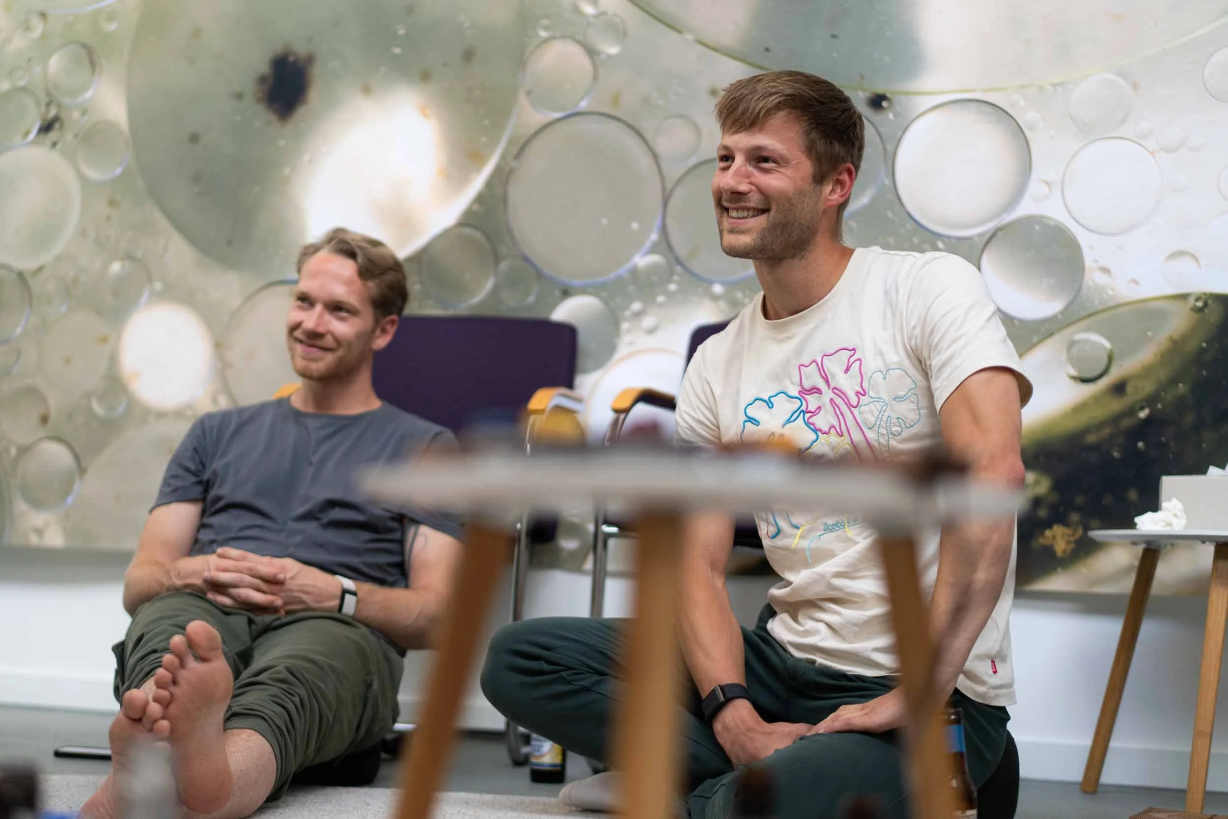 Two young men are sitting on the floor in a room with a large, abstract wall art featuring circular shapes in muted colors. One man is wearing a gray t-shirt and green pants, sitting barefoot with his legs extended and crossed at the ankles, smiling and looking to the side. The other man is wearing a white t-shirt with colorful outlines and dark pants, kneeling or squatting with his hands resting on his thighs, also smiling and looking in the same direction.
