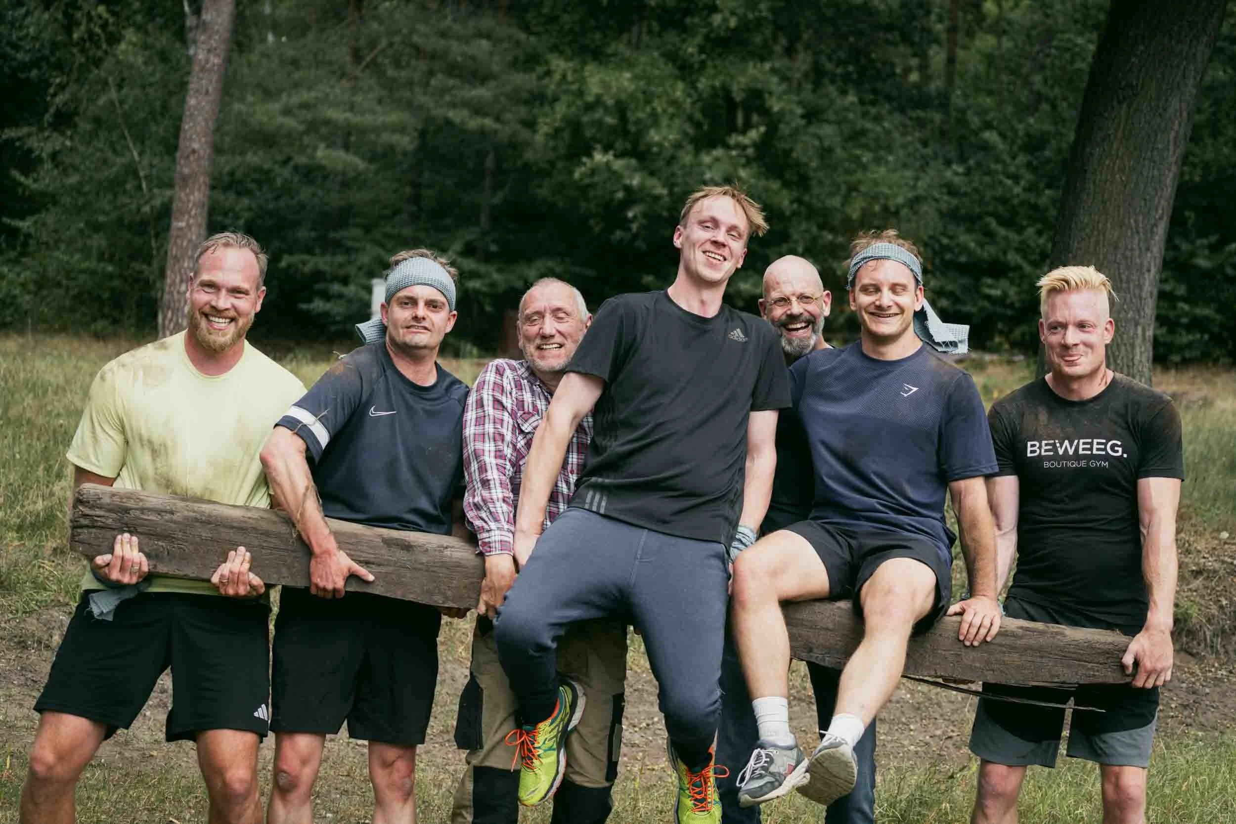 A group of seven men outdoors in a forest, posing for a photo. They are holding a large wooden log and appear to be in a playful or celebratory mood, with some smiling and others making humorous faces. Some are wearing workout clothes, and two are we