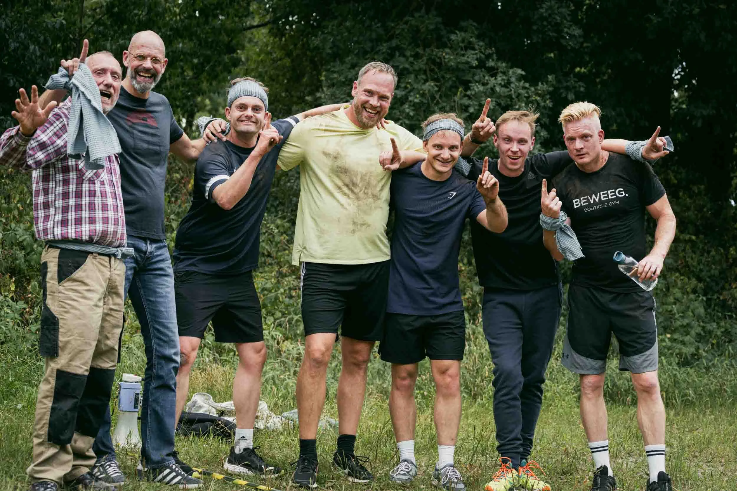 Group of seven men standing outdoors on grass, celebrating after a workout or running event, smiling, raising hands and pointing fingers, with trees in the background.