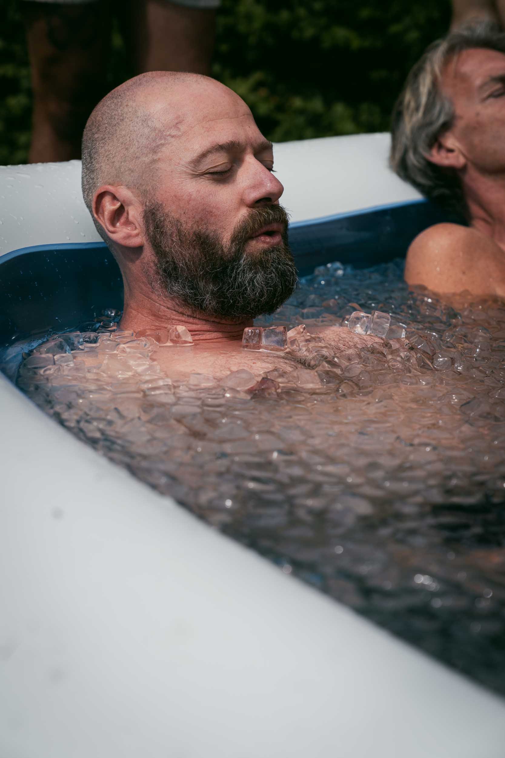 Two men relaxing in a hot tub filled with ice, with one man visible with his eyes closed and a beard, and another man partly visible with gray hair, outdoors with trees in the background.