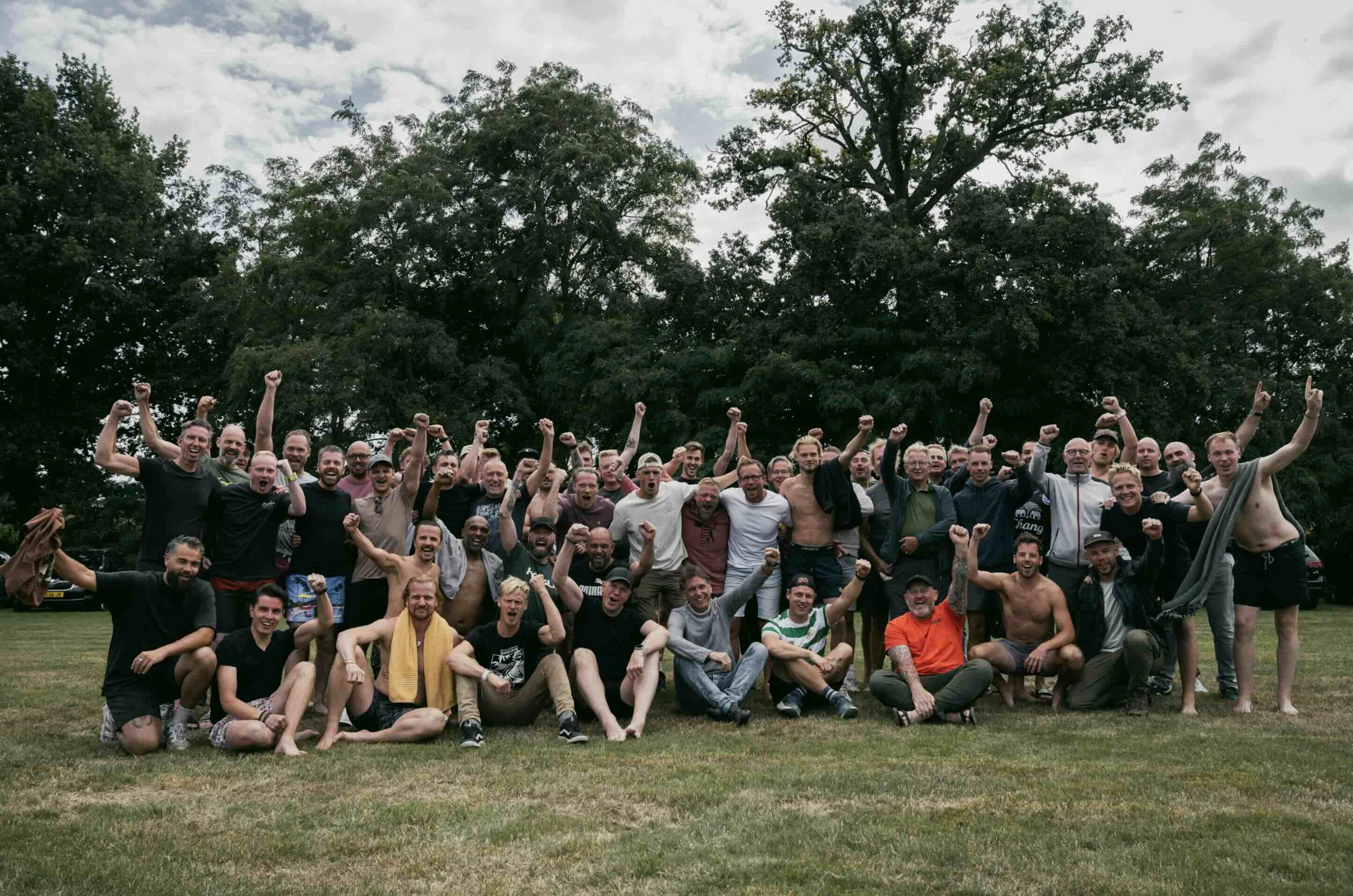 Large group of men gathered outdoors on a grassy field, celebrating with many raising their fists and cheering, with trees and cloudy sky in the background.