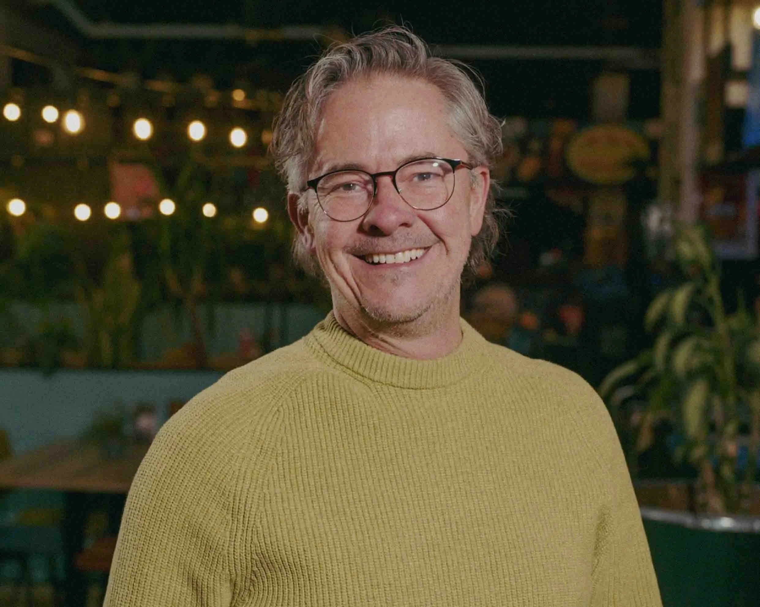 Group of men sitting outdoors, some with arms crossed, listening to a woman holding a beer bottle, with trees in the background.