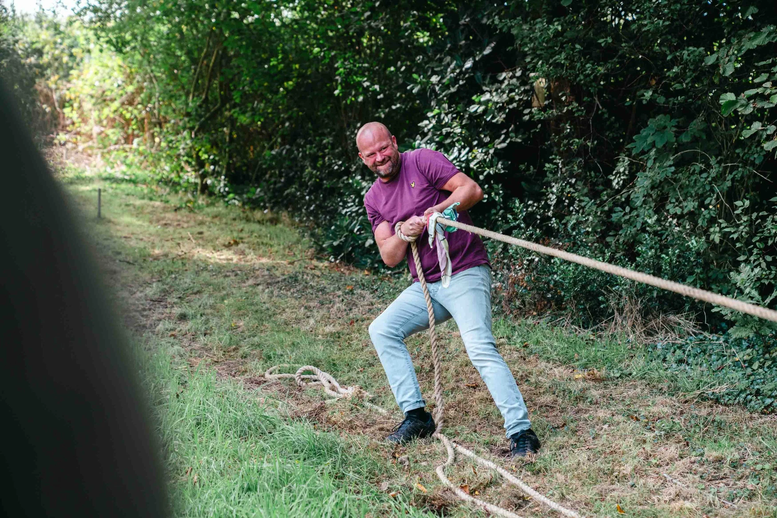 A man with a beard and a bald head pulling on a thick rope, smiling, in a grassy outdoor area with bushes and trees in the background.