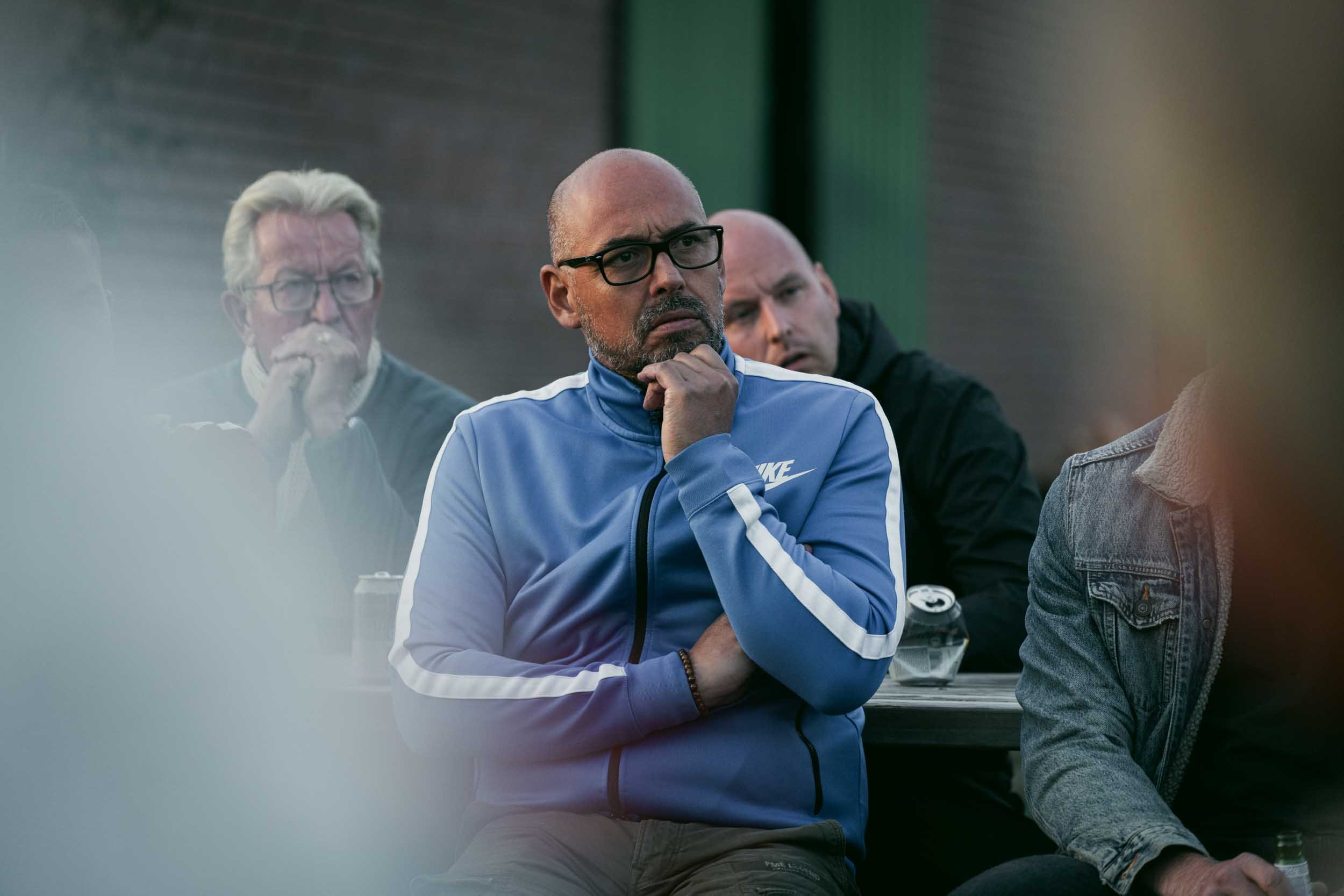 Four men seated at an outdoor table, with one man in focus wearing glasses and a blue Nike jacket, resting his chin on his hand with a pensive expression. The background is slightly blurred.