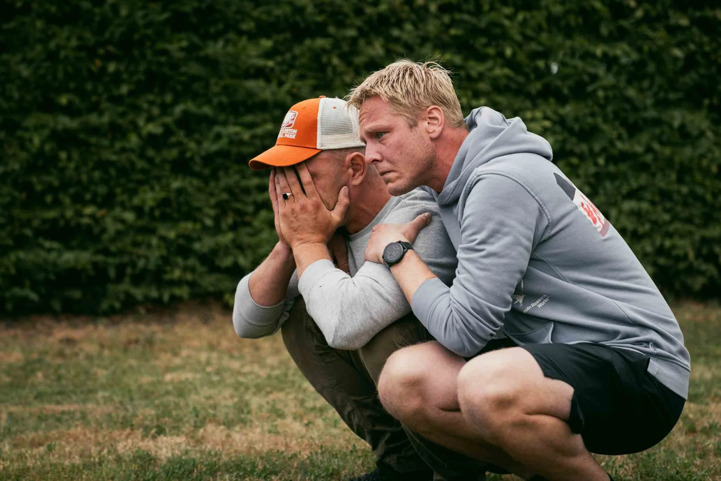 Two men crouching on the grass, one with his face covered by his hands and the other comforting him, both appear emotional, outdoors with a green hedge background.