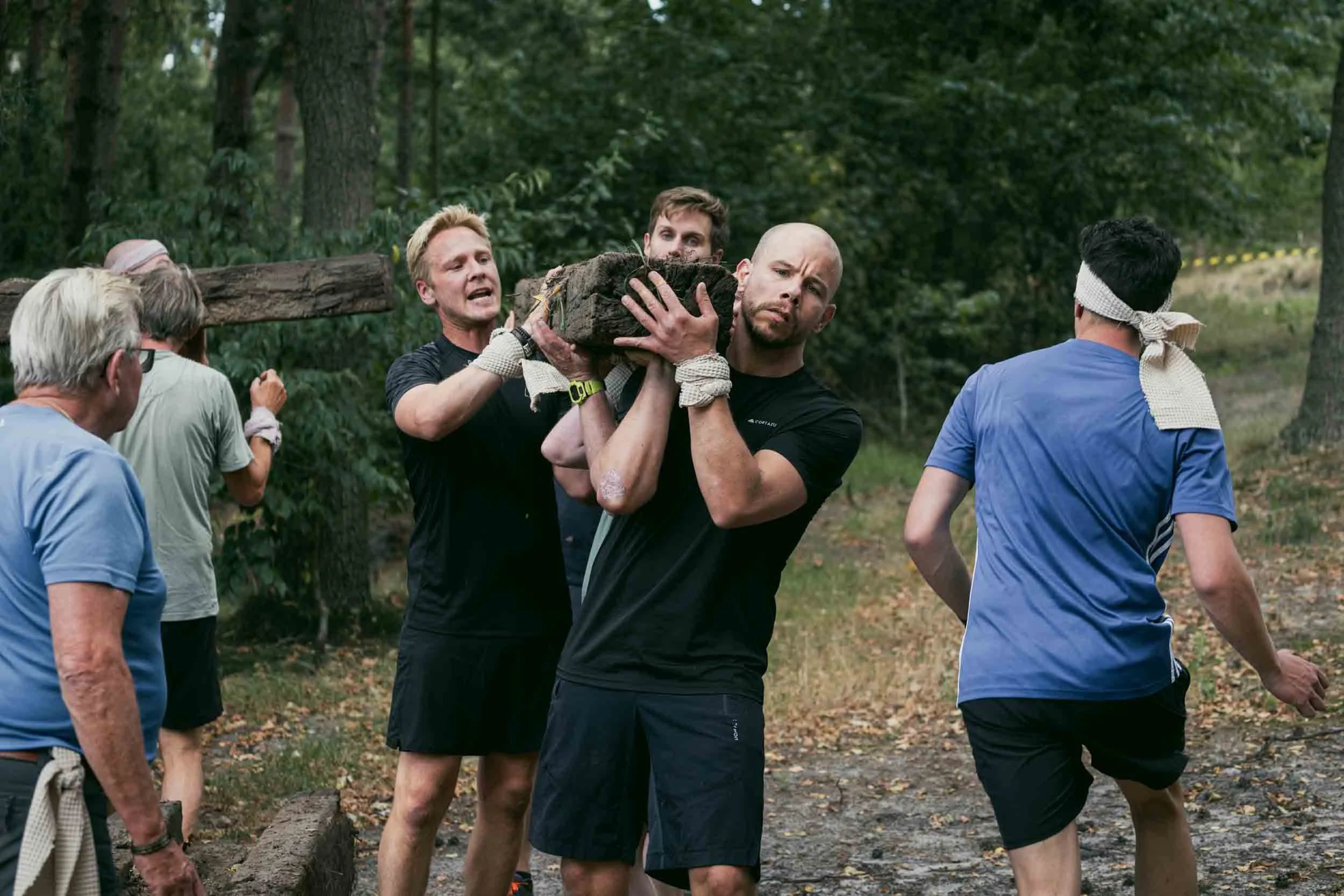 Group of people participating in a team-building activity outdoors in a wooded area, passing a large wooden log among them.